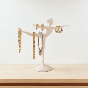 Könnte beinhalten: Ein weißer Schmuckhalter in Ballerina-Form, der verschiedene gold- und silberfarbene Armbänder, Ohrringe und eine Halskette zeigt. Der Schmuckhalter steht auf einer hellen Holzoberfläche vor einer weißen Wand.