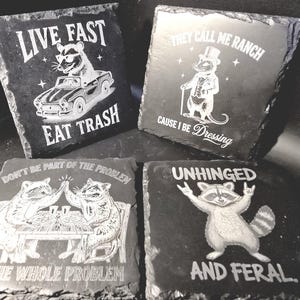 May include: Four square slate coasters with humorous raccoon designs. One coaster reads "Live Fast Eat Trash" with a raccoon driving a car. Another says "They Call Me Ranch Cause I Be Dressing". The third says "Don't Be Part of the Problem Be The Whole Problem". The last says "Unhinged And Feral."