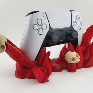 Peut inclure: Une manette de jeu blanche et noire repose sur un support rouge et dentelé avec deux crânes beiges. La manette a des boutons noirs et un centre noir. Le support a un aspect texturé et organique.