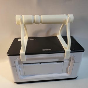 May include: A white and black printer with a white handle attached to the top. The handle is made of a light-coloured material and is designed to make the printer portable. The printer has the word "UNBYN" printed on the top.