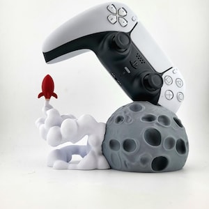 May include: A white and black PlayStation 5 controller rests on a gray moon-shaped base with a red rocket taking off. The base has a cloud design and is a unique gaming accessory.