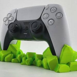 May include: A white and black PlayStation 5 controller displayed on a bright green, geometric stand. The stand is designed to resemble a cluster of stylized rocks, offering a unique display for the gaming accessory.
