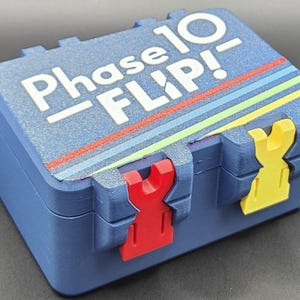 May include: A blue Phase 10 Flip game box with white text and colourful stripes. The box has red and yellow clasps. The game box is rectangular and has a textured surface.