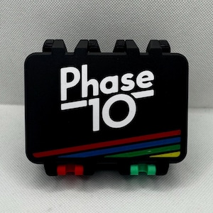 May include: Black Phase 10 card game case with white text and colourful stripes. The case has a rectangular shape with a hinged lid and small coloured tabs on the bottom. The text "Phase 10" is prominently displayed.