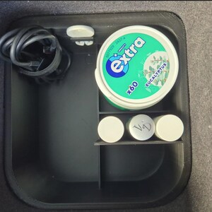 May include: A black storage compartment in a vehicle, containing a coiled black cable, a container of Extra Eucalyptus gum, and three small white containers. The gum container is green and white.