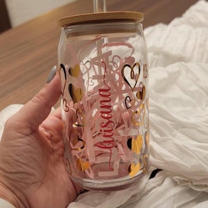 May include: A clear glass tumbler with a bamboo lid and a straw. The tumbler has a pink and gold design with hearts and the name "Luisaana" written on it.