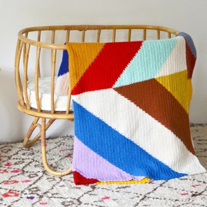 Op de afbeelding: Een geweven rieten wieg met een kleurrijk gehaakt deken eroverheen. Het deken is gemaakt van verschillende kleuren, waaronder rood, geel, blauw en groen.