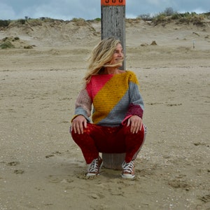 Könnte beinhalten: Eine Frau in einem bunten gestreiften Pullover und roten Hosen sitzt auf einem Holzpfosten mit einem Schild, auf dem "13 000" steht, an einem Sandstrand.