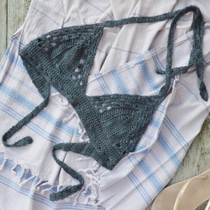 Op de afbeelding: Een donkergrijze gehaakte bikini top met halter stijl en een driehoekige vorm. De top ligt op een gestreepte handdoek.