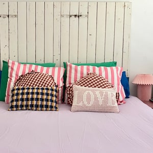 May include: A white wooden headboard with a pink, white, and red striped pillow, a red and white gingham pillow, a white crocheted pillow with the word "LOVE" embroidered on it, and a blue and yellow plaid pillow. The bed has a light purple sheet.
