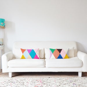 May include: A white sofa with two colorful geometric patterned throw pillows. The sofa is in a living room with a white rug, a white table with a lamp, and a white table with a candle holder.