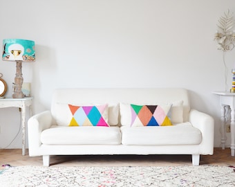 May include: A white sofa with two colorful geometric patterned throw pillows. The sofa is in a living room with a white rug, a white table with a lamp, and a white table with a candle holder.