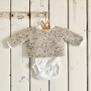 May include: A white onesie with a beige speckled knit sweater on top. The sweater is hanging on a wooden bunny-shaped hanger against a white wooden background.