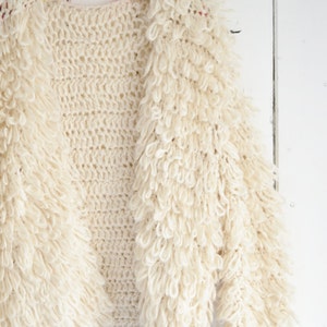 Puede incluir: Un cárdigan blanco de ganchillo con una textura esponjosa, colgado de una percha con una banda de rayas de colores. El cárdigan tiene un dobladillo con flecos.