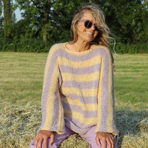 Op de afbeelding: Een vrouw in een geel-paarse gestreepte trui en lavendelkleurige broek zit op een hooibaal. Ze draagt een zonnebril en glimlacht.