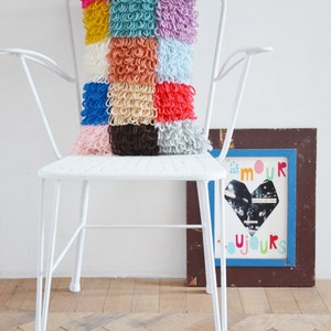 Può includere: Un cuscino colorato a patchwork con una varietà di trame e colori, tra cui rosa, blu, giallo e verde. Il cuscino è seduto su una sedia bianca con una struttura in metallo bianco.