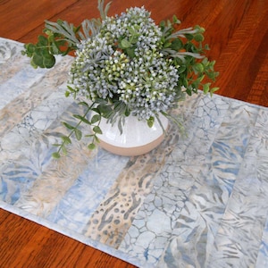 May include: A blue and beige patterned table runner with a floral design. A white and beige vase holds a bouquet of green and white flowers. The runner is on a wooden table.