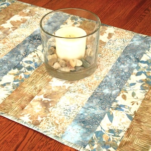 May include: A table runner with a blue and brown striped pattern. The runner is made of fabric and has a fringed edge. A glass candle holder with a white candle and small stones is on top of the runner.