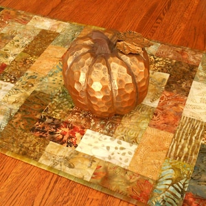 May include: A rectangular fabric table runner featuring a patchwork design of various earth-toned squares in shades of brown, tan, green, and cream, topped with a metallic copper-colored decorative pumpkin with a textured, hammered surface.