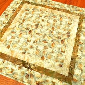 May include: A square fabric quilt featuring a central panel with a dense pattern of brown, tan, and sage green leaves. The center is framed by a solid dark olive green border and an outer border of the same leaf print, creating a layered design.