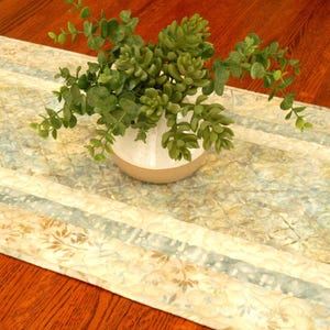 May include: A rectangular table runner featuring a marbled pattern in shades of light blue, cream, and tan with a subtle floral motif. A white ceramic vase containing a green artificial succulent plant sits centered on the fabric runner.
