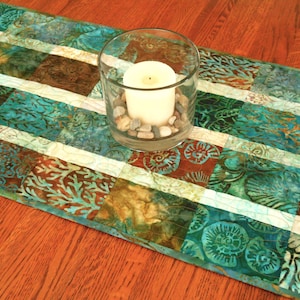 May include: A decorative table runner with a patchwork design in shades of teal, brown, and cream. A clear glass candle holder with a white candle and small stones sits on the runner. The runner is approximately 12 inches wide.