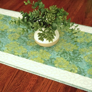 May include: A floral table runner with a light blue and green pattern. The runner has a white border with a quilted design. A small white and tan vase with green plants sits on top of the runner.