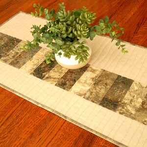 May include: A rectangular quilted table runner featuring a central band of alternating cream-colored fabric and marble-patterned blocks in shades of brown, gray, and white. A white ceramic vase containing a green faux succulent plant sits on top.