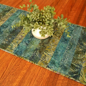 May include: A teal and olive green batik table runner with a leaf pattern. The runner is decorated with a white ceramic bowl filled with green plants. The runner is placed on a wooden table.