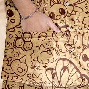 K&ouml;nnte beinhalten: Beige Hemd mit Kn&ouml;pfen und einem braunen Cartoon-Muster. Das Hemd hat eine Tasche und kleine wei&szlig;e Kn&ouml;pfe. Das Design umfasst Cartoon-Tiere, Blumen und andere dekorative Elemente.