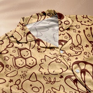 K&ouml;nnte beinhalten: Beigefarbenes Hemd mit Kn&ouml;pfen und einem sich wiederholenden Muster aus braunen Cartoon-Illustrationen, darunter Bienen, Hasen und Blumen. Das Hemd hat einen klassischen Kragen und wei&szlig;e Kn&ouml;pfe. Der Stoff wirkt weich und leicht.