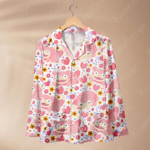 K&ouml;nnte beinhalten: Lang&auml;rmliges Pyjama-Oberteil mit wei&szlig;em Hintergrund und einem sich wiederholenden Muster aus rosa Cartoonfiguren, Herzen und Blumen. Das Pyjama-Oberteil hat einen Kragen, eine Knopfleiste und zwei Vordertaschen.