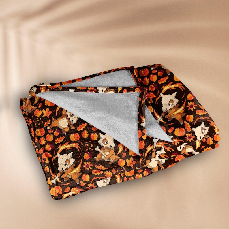 Op de afbeelding: Een opgevouwen deken met een herhalend patroon van stripfiguren, pompoenen en herfstbladeren in bruin, oranje en wit. De deken heeft een zachte, grijze binnenkant.