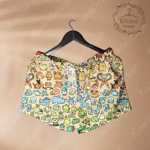 May include: Tan shorts with a colorful cartoon character pattern, hanging on a black hanger. The shorts have a drawstring waist and are decorated with various creatures in shades of blue, green, orange, and yellow.