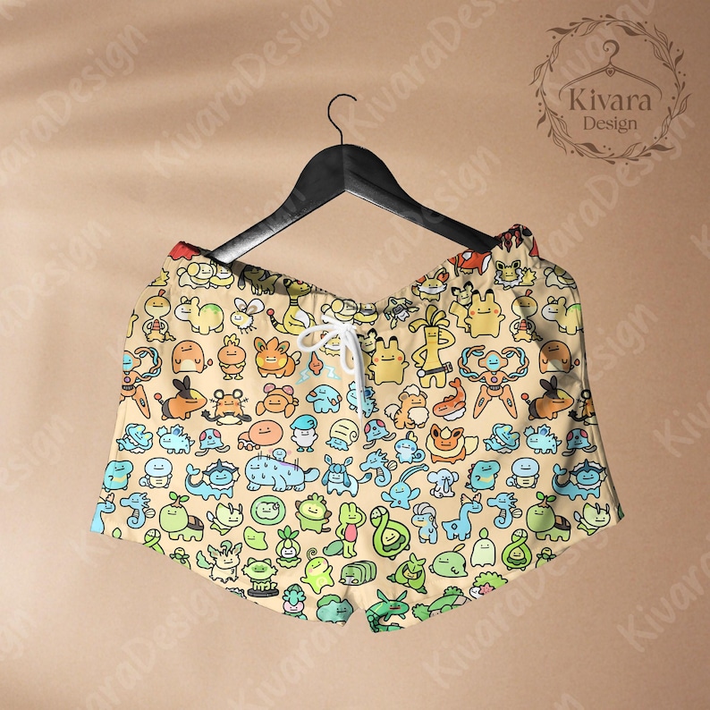 May include: Tan shorts with a colorful cartoon character pattern, hanging on a black hanger. The shorts have a drawstring waist and are decorated with various creatures in shades of blue, green, orange, and yellow.