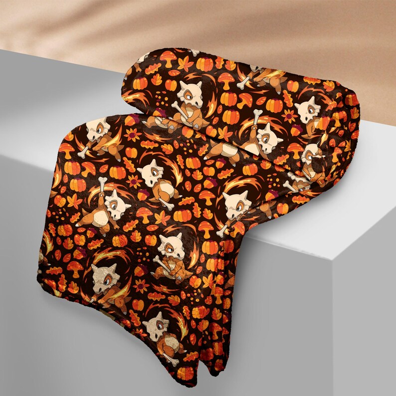 Op de afbeelding: Een donkerbruine deken met een herhalend patroon van oranje pompoenen, paddenstoelen en gestileerde Cubone-personages. De deken heeft een zachte, pluche uitstraling, met een gevouwen rand. Het ontwerp bevat herfstkleuren en thema's.