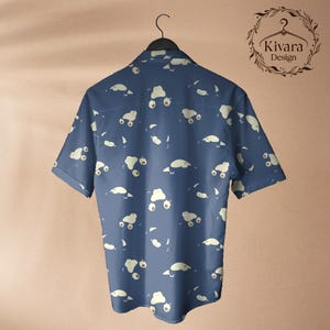 May include: A short-sleeved button-down shirt in a dark blue colour featuring an all-over repeating pattern of small, cream-coloured cartoon creatures. The shirt is displayed on a black hanger, and the upper right corner displays the text Kivara Design.
