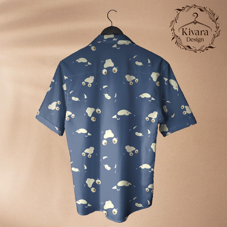 May include: A short-sleeved button-down shirt in a dark blue colour featuring an all-over repeating pattern of small, cream-coloured cartoon creatures. The shirt is displayed on a black hanger, and the upper right corner displays the text Kivara Design.