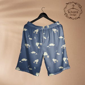 May include: Blue shorts featuring an all-over pattern of small, cream-coloured, rounded cartoon creatures. The shorts have an elastic waistband with a white drawstring and are displayed on a black hanger. The text Kivara Design is visible.