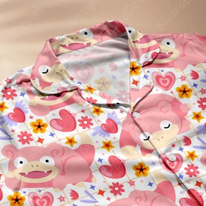 K&ouml;nnte beinhalten: Rosa Pyjama-Oberteil mit einem sich wiederholenden Muster aus Zeichentrickfiguren, Herzen, Blumen und Sternen. Das Pyjama-Oberteil hat einen Kragen und eine Knopfleiste vorne. Der Hintergrund ist wei&szlig; mit einem Gittermuster.