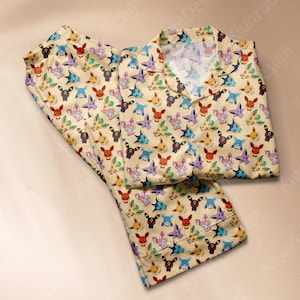 Op de afbeelding: Een beige pyjamaset met een herhaald patroon van kleurrijke stripfiguren. De set bevat een overhemd met kraag en knopen en een bijpassende broek. De karakters zijn in verschillende kleuren, waaronder rood, blauw, paars en groen.