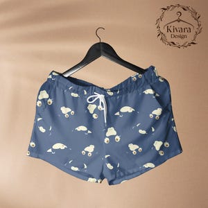 May include: Blue shorts featuring a repeating pattern of small, cream-coloured cartoon creatures with rounded shapes and visible paws. The shorts have an elastic waistband with white drawstring cords and are displayed on a black plastic hanger.