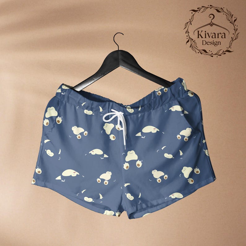 May include: Blue shorts featuring a repeating pattern of small, cream-coloured cartoon creatures with rounded shapes and visible paws. The shorts have an elastic waistband with white drawstring cords and are displayed on a black plastic hanger.