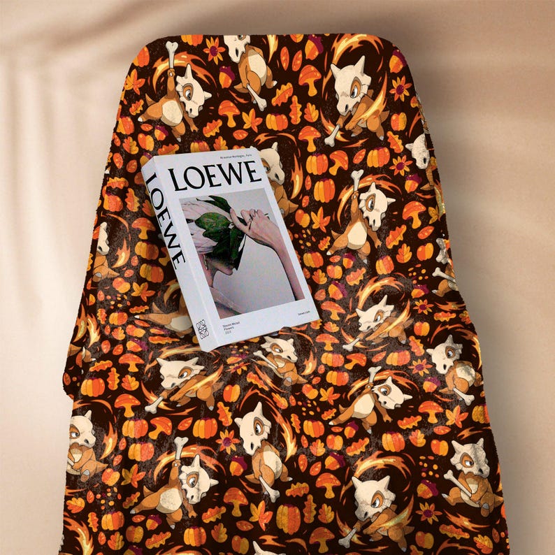 Op de afbeelding: Een bruine deken met een herhaald patroon van stripfiguren, pompoenen en herfstbladeren. Een wit boek met het woord "LOEWE" ligt op de deken.