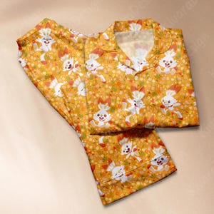 May include: A two-piece pajama set featuring a repeating pattern of white cartoon bunnies with orange accents on a yellow background. The pajamas include a collared button-down shirt and matching pants.