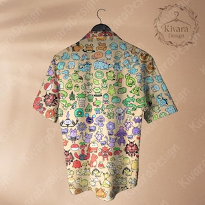 May include: A short-sleeved button-down shirt with a beige background and a colorful pattern of cartoon characters. The shirt is hanging on a black hanger. The shirt's design includes various creatures in shades of green, blue, red, and purple.