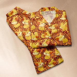 May include: A two-piece pajama set featuring a repeating pattern of cartoon foxes and flowers in shades of yellow, orange, and brown. The pajama top has a collared neckline and button closure. The fabric appears to be soft and comfortable.