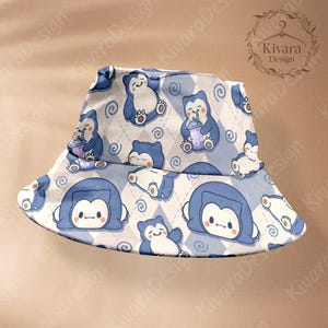 Puede incluir: Un sombrero de pescador azul y blanco con un patrón repetido de un personaje de dibujos animados. El patrón incluye al personaje en varias poses, algunos sosteniendo una bebida, sobre un fondo de diamantes y remolinos. El sombrero tiene un ala ancha.