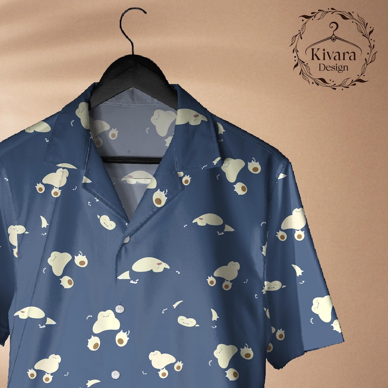 May include: A short-sleeved button-down shirt in dark blue featuring an all-over pattern of small, light-coloured, blob-like creatures. The shirt has a notched collar and is displayed on a black hanger. The logo Kivara Design is visible.