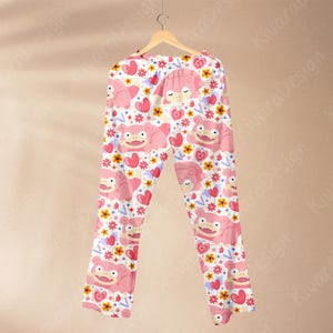 K&ouml;nnte beinhalten: Rosa Schlafanzughose mit einem sich wiederholenden Muster aus Zeichentrickfiguren, Herzen und Blumen. Die Hose h&auml;ngt an einem Holzkleiderb&uuml;gel vor einem neutralen Hintergrund. Die Schlafanzughose ist aus einem weichen, leichten Stoff gefertigt.